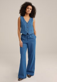 Modra jeansa brezrokavnik z globokim V-izrezom in tremi gumbi, v kombinaciji z ohlapnimi modrimi hlačami z elastiko in vrvico za zategovanje.