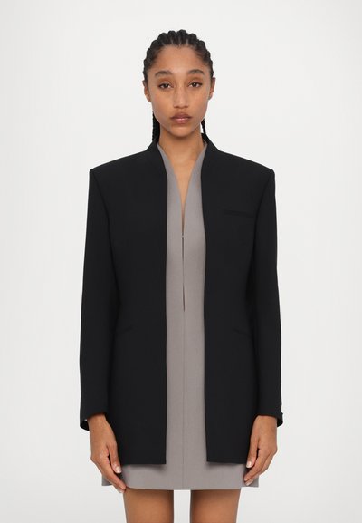 Černý oblekový blazer s vysokým límcem, úzkým střihem a bez knoflíků; nošený přes šedé šaty. Hladká textura, minimalistický design, boční kapsy.