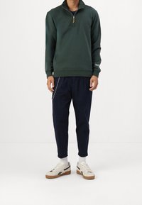 Sweatshirt vert à col zippé en tissu doux, associé à un pantalon marine avec un détail en chaîne. Baskets blanches avec des accents noirs.