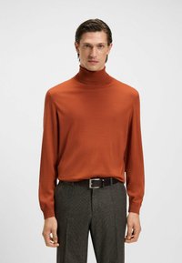 Ruda turtlenecko megztinis su ilgomis rankovėmis, raukšlėtomis rankogaliais, minkšta tekstūra. Suporuotas su pilkomis patikrintomis kelnėmis ir juodu diržu.