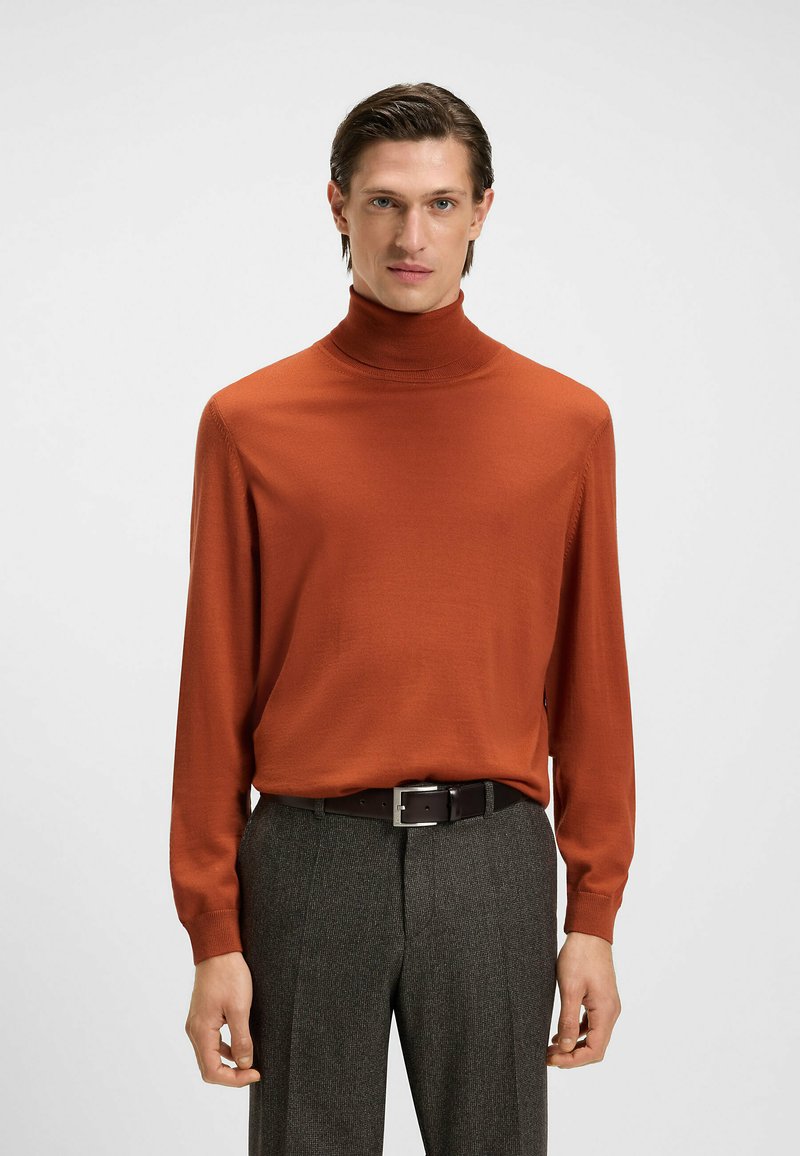 Ruda turtlenecko megztinis su ilgomis rankovėmis, raukšlėtomis rankogaliais, minkšta tekstūra. Suporuotas su pilkomis patikrintomis kelnėmis ir juodu diržu.