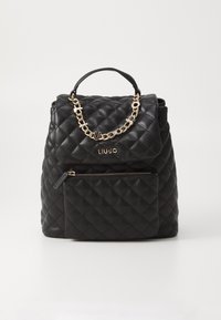 Mochila negra acolchada con un acento de cadena dorada, asa superior y bolsillo con cremallera frontal, con una textura de cuero suave. Nombre de la marca visible.