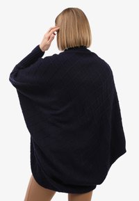 Lühikeste blonde juustega inimene, kes kannab tumedast sinisest tekstuuriga kudumit, seisab seljaga, käsi puudutades oma pead.