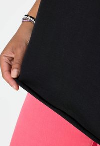 Mano con pulseras de cuentas sosteniendo el dobladillo de una camisa negra sobre pantalones rosas contra un fondo blanco.