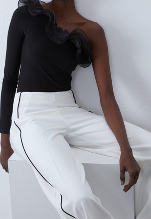 Vrouw draagt een zwarte top met één schouder en ruches en een witte high waist broek met zwarte bies, zittend op een witte blok.