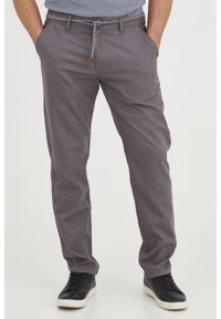 Man i grå avslappnade byxor med ett repbälte och svarta sneakers, stående med händerna i fickorna mot en neutral bakgrund.