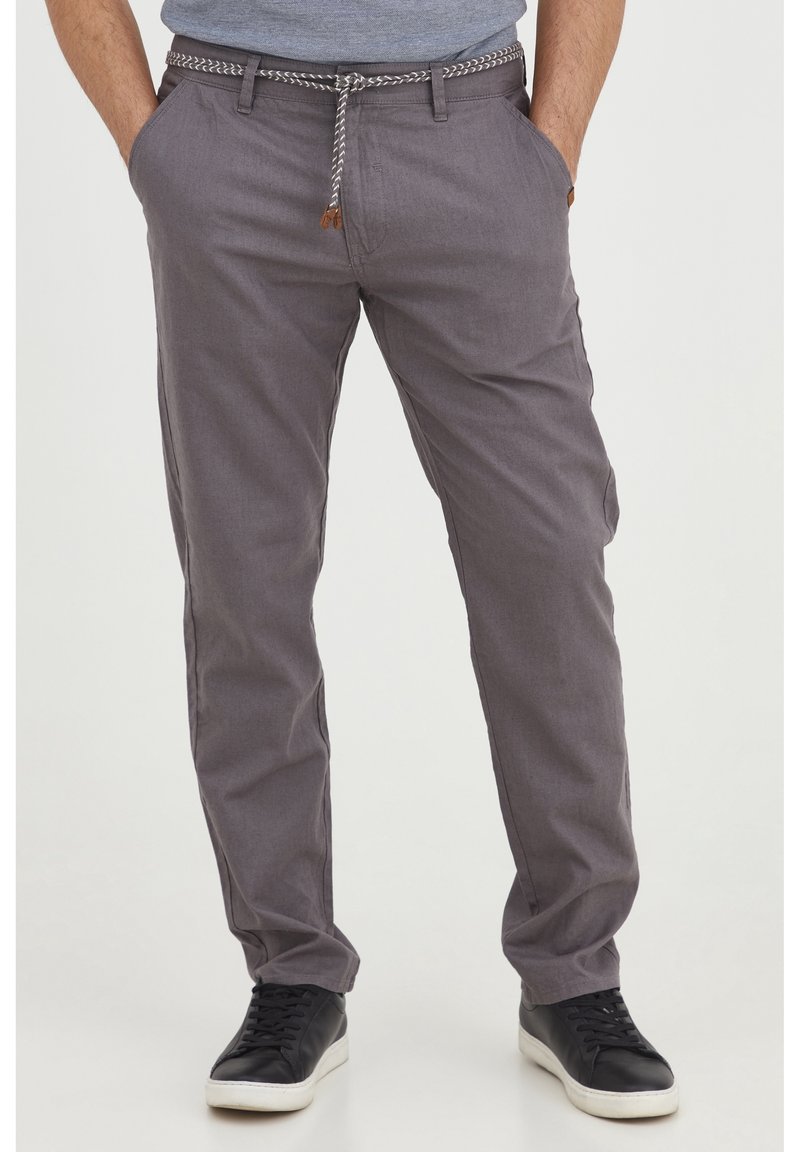 Man i grå avslappnade byxor med ett repbälte och svarta sneakers, stående med händerna i fickorna mot en neutral bakgrund.