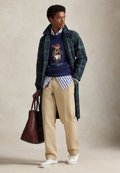 Mann iført rutete lang frakk, marineblå genser med bjørnegrafikk, stripete skjorte, beige bukser, hvite joggesko, holder stor brun skinnveske, står og ser mot venstre.
