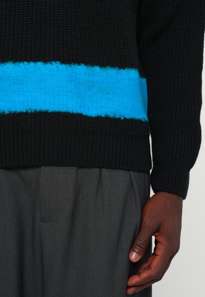 Schwarzer Strickpullover mit einem strukturierten blauen Streifen über der Brust. Kombiniert mit grauen gefalteten Hosen, die einen glatten Stoff zeigen.