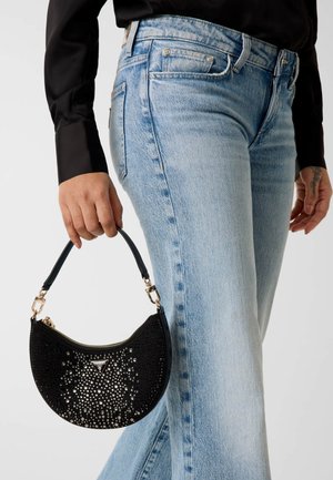 Persoon die lichtblauwe spijkerbroek en zwart shirt draagt, met een kleine zwarte halvemaanvormige handtas met zilveren studs en een bovenste ritssluiting.