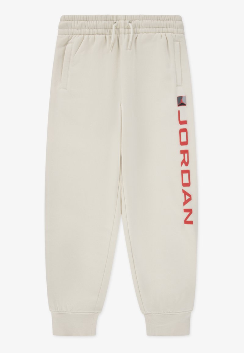 Μπεζ παντελόνι φόρμας με ελαστική μέση, πλαϊνές τσέπες και κόκκινο κείμενο "JORDAN" στο αριστερό πόδι. Μαλακό ύφασμα, στενά τελειώματα.