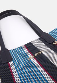 Vävd tygpåse med ett geometriskt mönster i blått, rött och vitt. Svarta konstläderhandtag och guld-logotyp på framsidan.