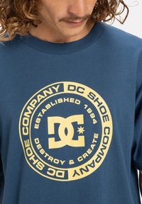 Modrá bavlněná tričko s velkým kulatým logem v žluté barvě, textem "DC SHOE COMPANY" a "DESTROY & CREATE", s kulatým výstřihem.