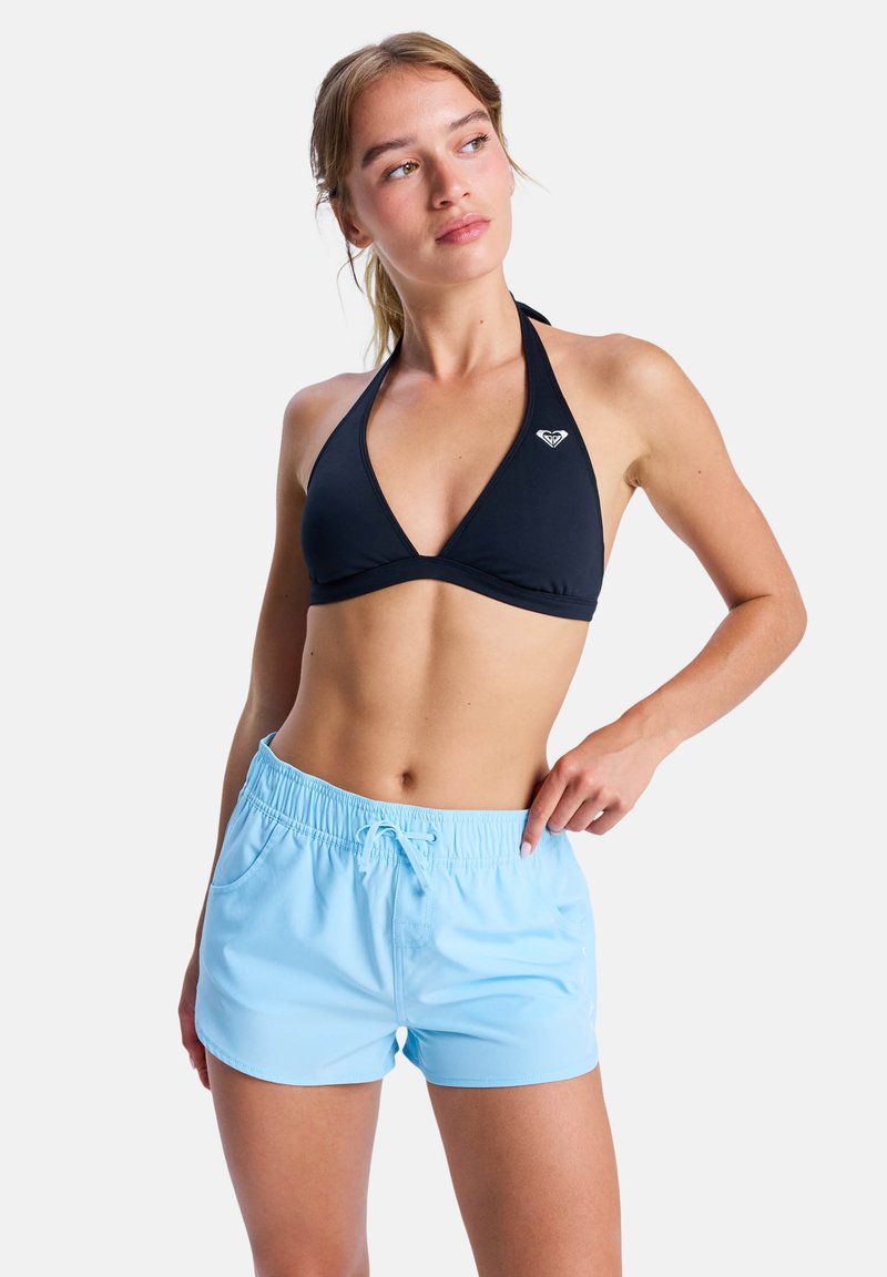 Young woman wearing a black halter bikini top and light blue swim shorts, looking to the side with one hand resting on the hip.