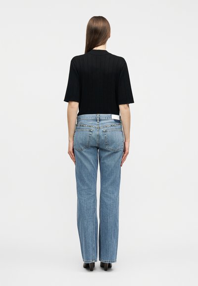 Γυναίκα με μακριά καστανά μαλλιά, ντυμένη με μαύρο κοντομάνικο μπλουζάκι και ψηλόμεσα ανοιχτό μπλε τζιν παντελόνια με ίσια γραμμή, στέκεται με την πλάτη γυρισμένη σε λευκό φόντο.