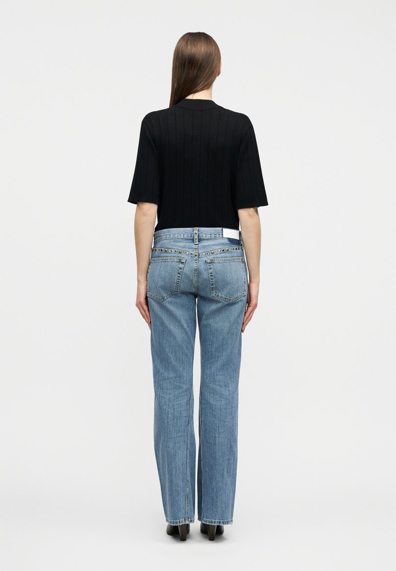 Kvinde med langt brunt hår iført sort kortærmet top og lyseblå højtaljede jeans med lige ben, stående med ryggen mod på hvid baggrund.