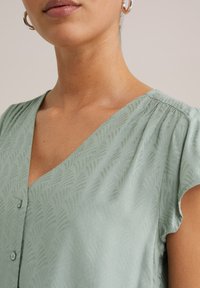 V-ringad, kortärmad blus i mjukt, texturerat mintgrönt tyg med subtilt bladmönster och knapppdetaljer.