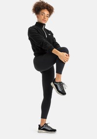Black fleece zip-neck pullover, fitted black leggings, and black shoes with gray accents. Model is demonstrating a leg stretch.