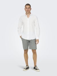 Chemise boutonnée blanche en lin à manches longues, assortie à un short gris et à des baskets noires. L'arrière-plan est d'un gris clair uni.