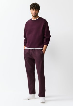 Un homme se tient debout, les mains dans les poches, portant un sweat-shirt violet foncé et un pantalon, des chaussettes blanches et une chemise blanche visible à la taille.