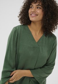 Top verde texturizado con escote en V y mangas sueltas. Presenta una tela suave y un detalle de costura sutil. Apto para uso casual.