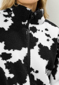 Veste en polaire noir et blanc avec un motif de vache, fermeture éclair complète et col haut. Texture douce, design décontracté.