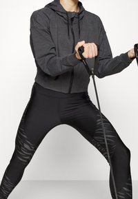 Mörkgrå kort hoodie med dragkedja, i kombination med svarta leggings med en strukturerad randig design på sidorna. Tränar med motståndsband.