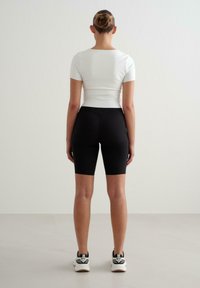 Bílý top s krátkým rukávem v kombinaci s černými cyklistickými šortkami, oba přiléhavé. Modelka má na sobě černé a bílé sportovní boty. Neutrální pozadí.