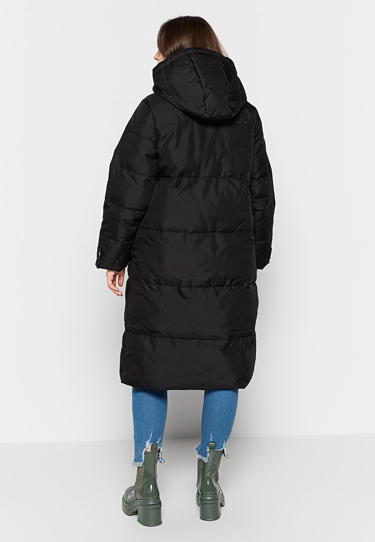 Femme portant un long manteau rembourré noir avec capuche, un jean skinny déchiré bleu et des bottines à talons vertes, de dos.