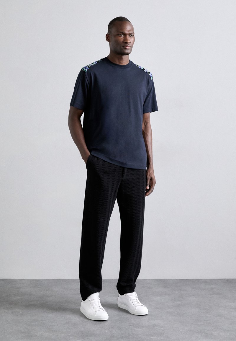 T-shirt bleu marine à manches courtes avec des accents de épaule à motifs, associé à un pantalon noir texturé et des baskets blanches. Coupe décontractée et design moderne.