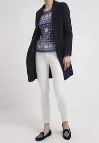 Marino kappa, mönstrad blå tröja, vita skinny jeans och marinblå broderade tofflor skapar en lager-på-lager, avslappnad outfit.