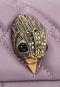 Pelle lavanda trapuntata con una spilla in tonalità oro che presenta un design di piuma dettagliato e accenti neri, evidenziando una texture ornamentale.