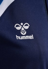 Donkerblauwe stof met een gladde textuur, met daarop een wit hummel-logo, inclusief een embleem van een bij en de merknaam eronder.