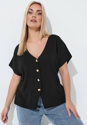 Lyshåret kvinde iført en sort V-hals knapbluse og blå jeans, poserer mod en hvid væg med et neutralt udtryk.