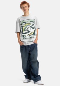 Szara, oversize'owa koszulka z graficznym wzorem przedstawiającym sneaker i napisem "DC Athletics"; zestawiona z luźnymi, ciemnoniebieskimi dżinsami i białymi sneakersami.