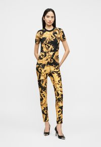 Γυναίκα στέκεται με τα χέρια πίσω από την πλάτη, φορώντας ένα μαύρο και χρυσό σετ με μπαρόκ τύπωμα, στενό τοπ με κοντά μανίκια και παντελόνι, συνδυασμένο με μαύρες γόβες.