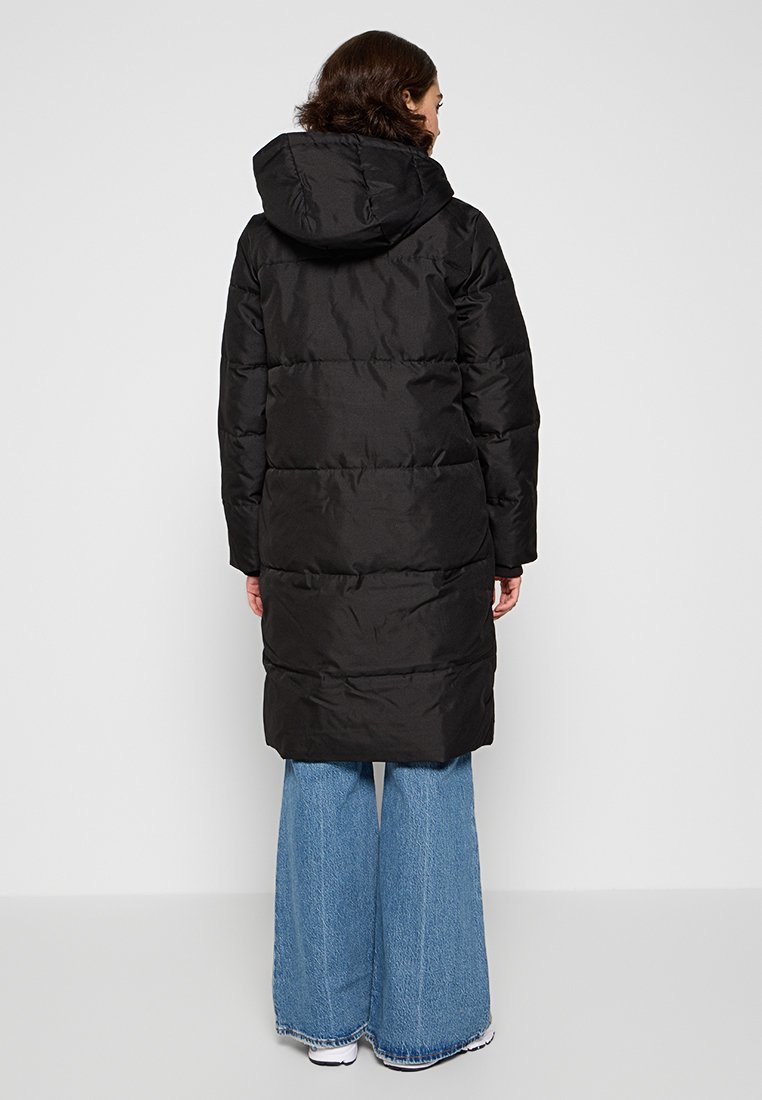 Schwarze Daunenjacke mit Kapuze, mit horizontalen Nähten, längerer Länge und glatter, glänzender Außenseite. Kombiniert mit weiter geschnittenen Jeans.