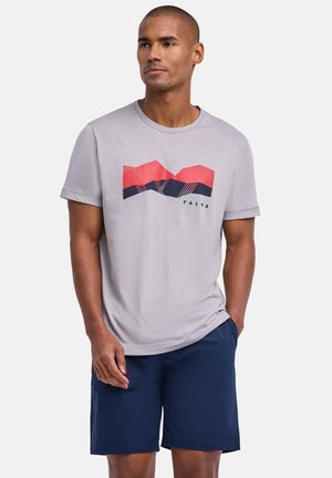 Homme portant un t-shirt gris clair avec un motif rouge et bleu marine et un short bleu marine, debout avec une main dans la poche du short, regardant vers la droite.