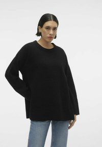 Maglione nero a maglia con una vestibilità comoda, maniche larghe e scollatura rotonda, abbinato a jeans azzurri su uno sfondo semplice.