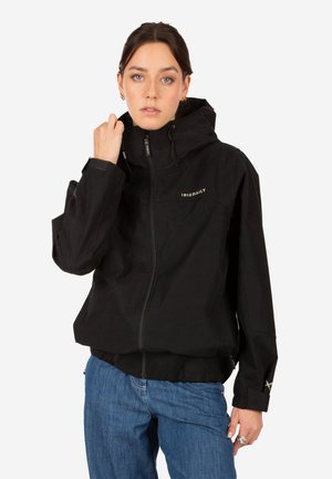 Woman wearing a black IRIEDAILY hooded jacket and blue jeans, holding the jacket collar with one hand.