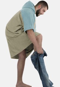 Kapuze-Bademantel in Beige und Hellblau, aus weichem Frottee. Eine Person zieht sich barfuß blaue Jeans an.