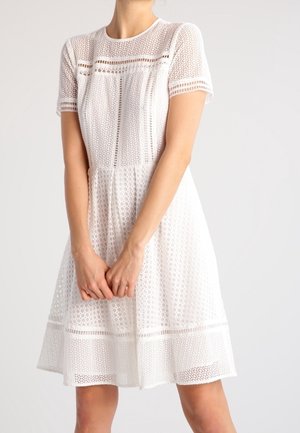 Woman wearing a knee-length white lace dress with short sleeves and geometric openwork patterns on a light background.