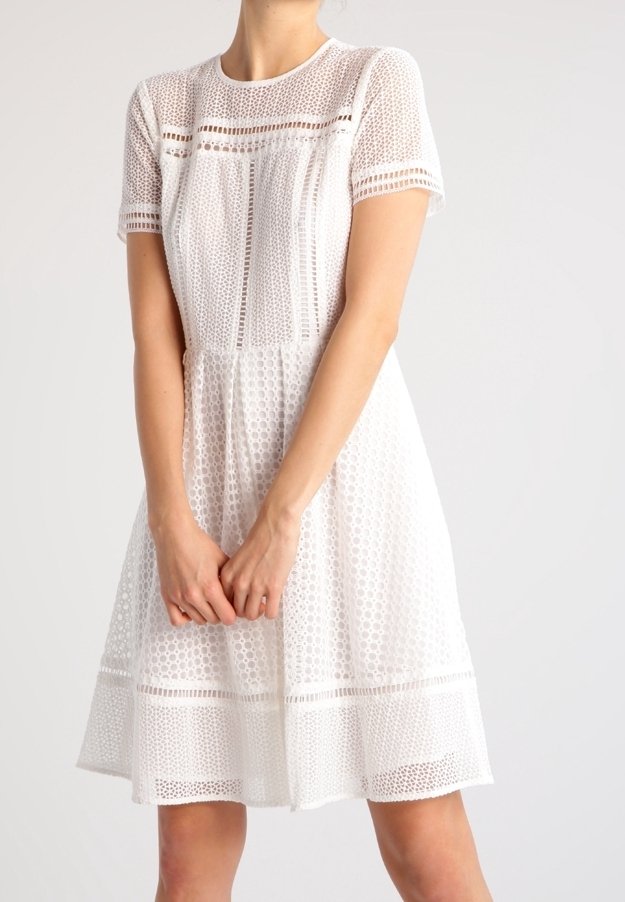 Femme portant une robe en dentelle blanche au genou avec des manches courtes et des motifs géométriques en ajour sur un fond clair.