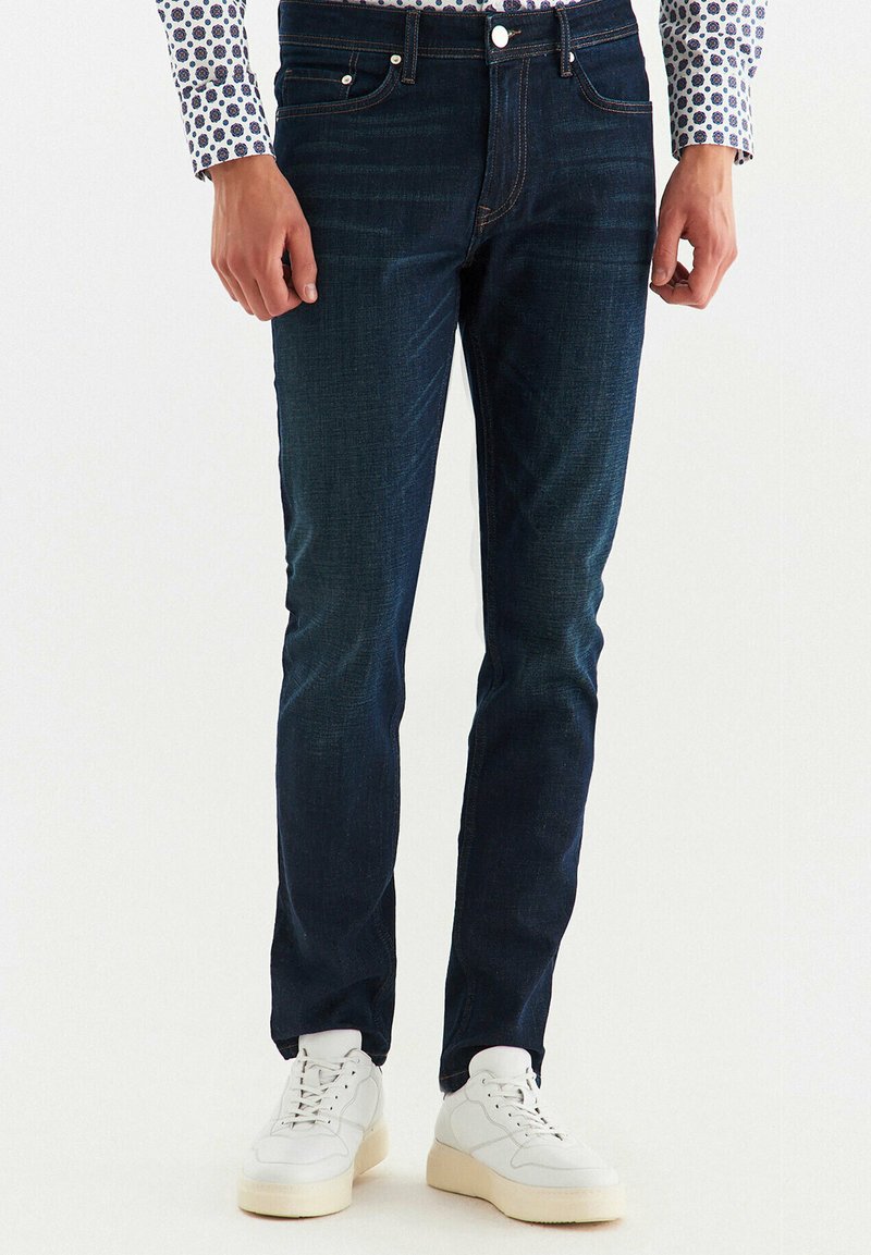 Granatowe dżinsy o slim fit z lekkim przetarciem, pięcioma kieszeniami i kontrastowym przeszyciem. W połączeniu z białymi sneakersami.
