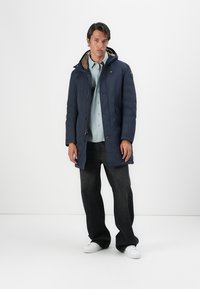Cappotto con cappuccio color navy, con un'esterno texturizzato, chiusura con zip, tasche laterali e un colletto foderato in marrone. Indossato sopra una camicia azzurro chiaro e jeans scuri.