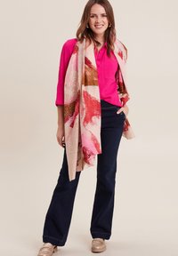 Sciarpa con motivo floreale nei toni del rosa, arancione e oro, lasciata cadere su una maglietta rosa brillante e strutturata. La modella indossa pantaloni svasati scuri e scarpe beige.