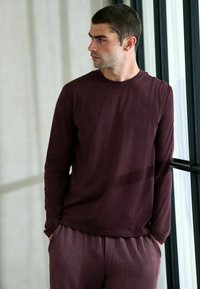 Bordeauxrotes Langarmshirt kombiniert mit gestreiften bordeauxroten Hosen. Der Stoff wirkt weich und leicht, mit lockerer Passform und rundem Ausschnitt.