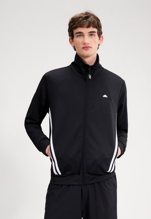 Joven con cabello corto y castaño que lleva una chaqueta negra con cremallera y franjas blancas a los lados, de pie frente a un fondo liso y claro.