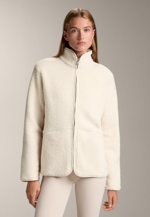 Bijela flis jakna s visokim ovratnikom, prednjim patentnim zatvaračem i dva velika džepa; mekani materijal i opušteni kroj.