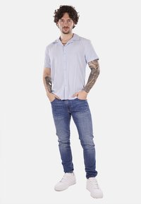 Camicia azzurra chiara a maniche corte, slim fit, indossata con jeans blu scuro e sneaker bianche. Il modello ha dei tatuaggi e sta in piedi con le mani nelle tasche.